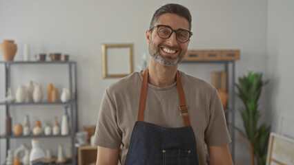 Man smiling in denim apron among ceramic pots on shelves in studio wearing glasses and beard, front...