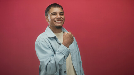 Man smiling and laughing while pointing, raising a clenched fist near his chest and touching his face in a studio with red backdrop; playful joy.