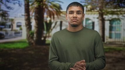 Fototapeta premium Man with hands clasped and a wry grimace, wearing an olive long sleeve shirt, standing near palm trees in front of a sunlit building; skepticism.
