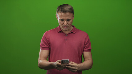 Man in red polo holding smartphone with both hands looking down and typing with thumbs in studio; surprise.