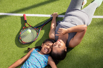 Asian mother and daughter relaxing on tennis court by white line with two rackets, ball