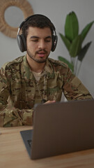 Fototapeta premium Man in military uniform listening with headphones at laptop in a cozy modern living room interior; focus.