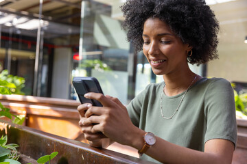African american woman checking smartphone on planter bench by glass wall with plants in cafe