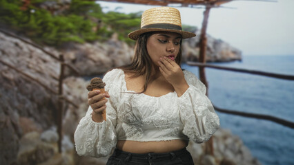 Woman holding icecream cone in hands with a slight pout near a rustic building railing on a cliff...