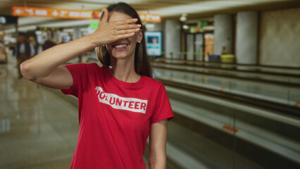 Woman covers eyes with hand in airport terminal wearing red volunteer tshirt and smiling; joy welcome volunteering.