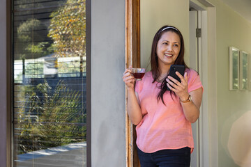 Asian woman holding mobile while leaning against door frame at home, sipping from glass cup