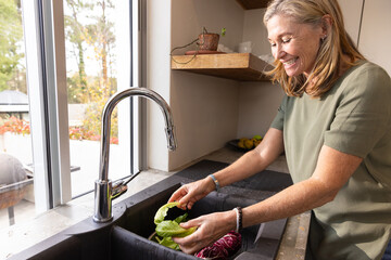 Senior woman washing lettuce and radicchio under chrome faucet at sink by window