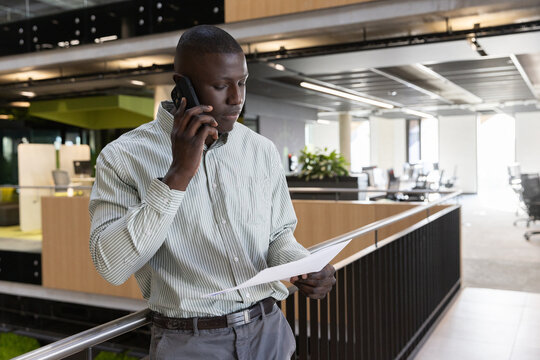 African american man in shirt with smartphone, reviewing document on office mezzanine railing - Powered by Adobe