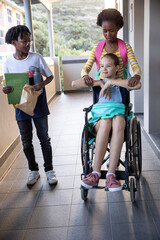 Diverse classmates pushing wheelchair walking down corridor holding folder bag water bottle
