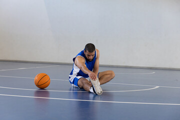 Male basketball player in blue uniform sitting stretching leg on basketball court near ball