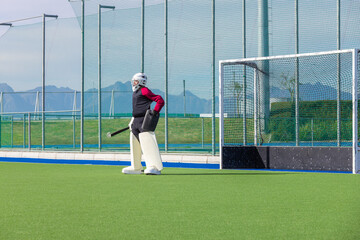 Male field hockey goalkeeper in protective gear guarding goal on synthetic turf holding stick
