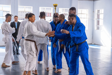 Diverse male martial artists gathering in dojo, holding gold trophy while in blue white gis