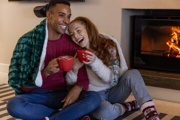 Naklejka premium Diverse couple sitting in living room wrapped in green fleece blanket with red mugs by fireplace