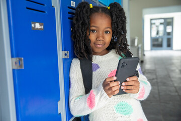African american child with smartphone and leaning against lockers with backpack in school hall