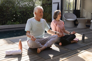Obraz premium Senior couple meditating at backyard deck beside pool with yoga mats, towel and water bottles