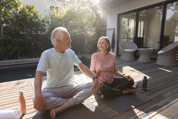 Senior couple stretching arms outward on yoga mats by pool wearing fitness trackers