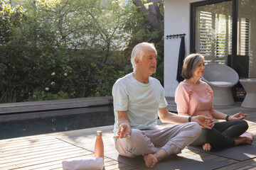 Senior couple meditating cross-legged on yoga mats at backyard deck beside pool with water bottle