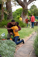 African american family hugging near brick-edged flower bed, using watering can in garden