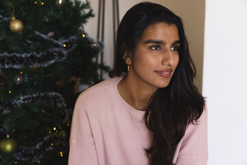 South Asian woman sitting by decorated Christmas tree at home wearing pink sweater gazing right