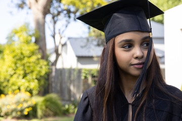 Female graduate standing in backyard wearing black gown and mortarboard by fence, copy space