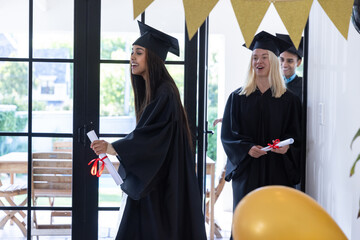 Obraz premium Diverse classmates in gowns walking through doors at home holding diploma scrolls, gold balloon