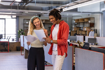 Diverse coworkers reviewing printed documents in modern office with desktop computers and shelving