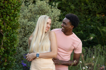 Fototapeta premium Diverse couple embracing each other, showcasing smartwatch along narrow garden path