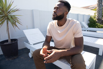 African American man sitting on slatted lounge chair near black planter on patio gazing upward