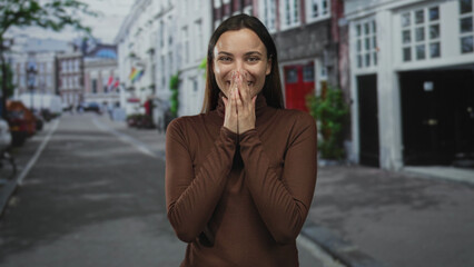 Obraz premium Woman with hands clasped near face smiling broadly while wearing brown turtleneck on a street lined with buildings; simple joy.
