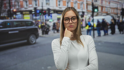 Obraz premium Young woman with hand on chin standing at a busy urban street crossing near traffic and pedestrians; thoughtful reflection.
