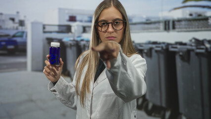 Fototapeta premium Woman holding a blue bottle and presenting it with an outstretched hand on a street with trash bins and parked car; confidence.