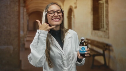 Young woman in lab coat holding a medicine bottle and making a call me hand gesture in a building corridor; playful promotion.