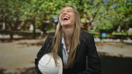Obraz premium Woman in business suit holding white hardhat laughing with mouth open at a city park with trees and benches; career confidence.