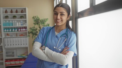 Hispanic female nurse smiling in a hospital room with arms crossed, wearing blue scrubs and stethoscope, suggesting a professional healthcare environment indoors.