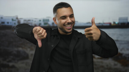 Man holds one thumb up and one thumb down gesture on a street near a rocky beachfront scene  indecision dilemma. © Krakenimages.com