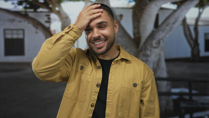Man smiling with hand to forehead on busy street in front of white building; relief recovery calm contentment.
