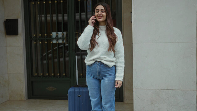 Young woman talking on phone with suitcase on street, wearing casual sweater and jeans, standing outdoors near entrance on a city sidewalk.