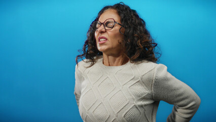 Woman experiencing pain, wearing glasses and sweater, standing against bright blue background,...