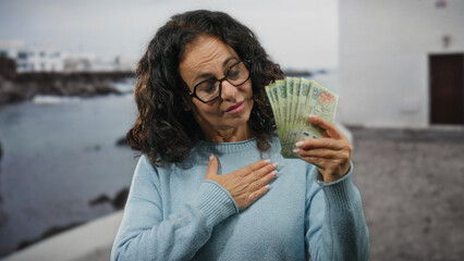 Middle-aged woman with curly hair holding argentine pesos outdoors by the seaside with a boat in the background, expressing emotion with hand on chest near a port.