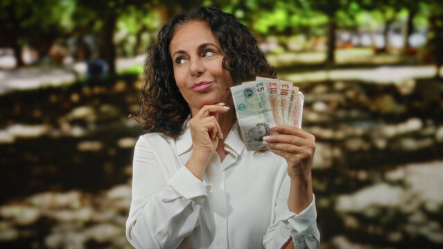 Woman holding british pounds thoughtfully outdoors in a park setting, conveying a sense of contemplation and decision-making with currency in hand under sunlight.