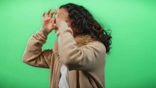 Woman with curly hair makes binoculars gesture against a green background, wearing a beige jacket, suggesting a playful and imaginative setting.