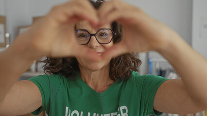Woman wearing green volunteer shirt making heart shape with hands indoors at charity room with...