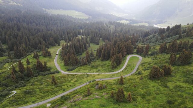 Mountain bikers riding downhill on a winding alpine road through forests and meadows in Switzerland. Aerial view of dynamic downhill cycling scene in the Swiss Alps.