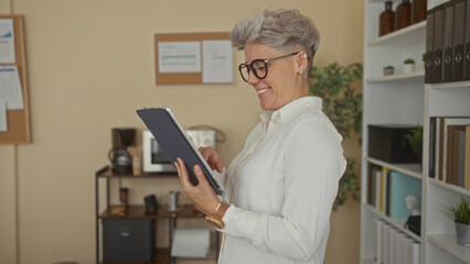 Woman touches tablet with finger in office lined with shelves and binders while smiling at screen; concentration.