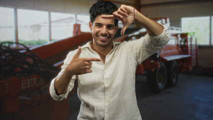 Obraz premium Man young hispanic framing the scene with hands and smiling, pointing finger gesture near his face in front of orange farm machinery inside a building; playful energy.