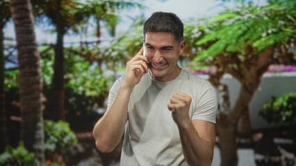 Man on phone making a fist pump with clenched hand in forest park outdoors, smiling and celebrating a win; triumphant excitement.