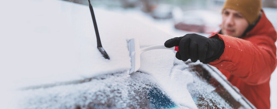 man in winter clothes clean ice and snow from car's windshield with scraper warming up car parked on residential street cold morning. Scene everyday winter life weather conditions and urban lifestyle