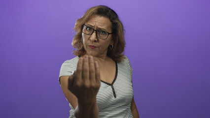 Middle aged hispanic woman pointing finger forward in purple studio; assertion invitation seriousness.