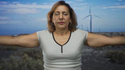 Middle aged hispanic woman lifts bare arms overhead in building exterior under bright blue sky; serenity.
