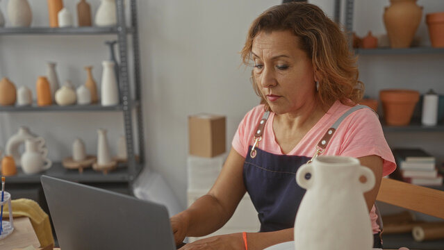 Artisan woman types on laptop beside handmade pottery vase and ceramic tools on wooden table in studio workspace, hands poised over keyboard; concentration. - Powered by Adobe